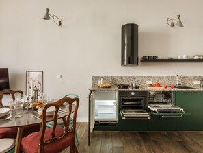 Una cocina moderna con horno empotrado, microondas y lavavajillas, una zona de comedor con sillas y una mesa para dos, y una lámpara colgante.