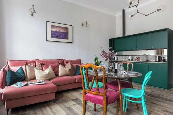 A modern living room with a sectional sofa, a dining table with chairs, and a kitchen area with green cabinets.