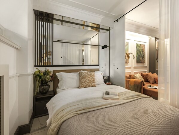 A modern bedroom with a large bed, a bedside table, a nightstand, a mirror, and a view into an adjacent living area.