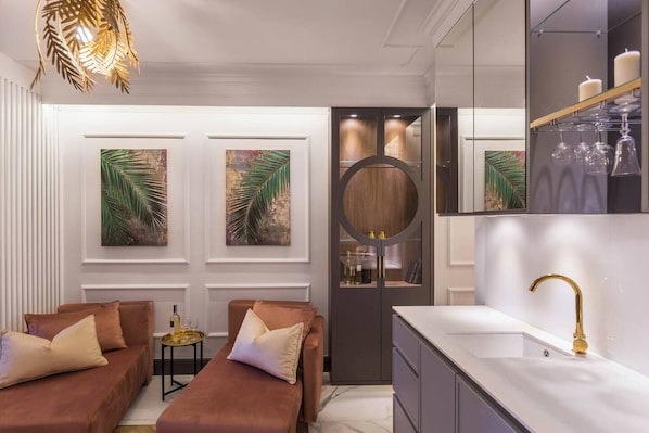 A modern bathroom with a white sink, gold faucet, and a round mirror. There are two brown sofas with cushions and a small round table with a bottle.