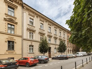 A street view with a row of parked cars, a building with classical architecture, and a tree on the sidewalk.