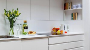 Une cuisine moderne avec des meubles blancs, une cuisinière encastrée, un évier et un plan de travail agrémenté d’un vase de feuilles vertes, d’une bouteille d’huile d’olive et d’un panier de fruits.