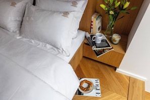 A neatly made bed with white linens and pillows, a wooden nightstand with a tray holding a mug, a book, and a candle, and a magazine on the floor.