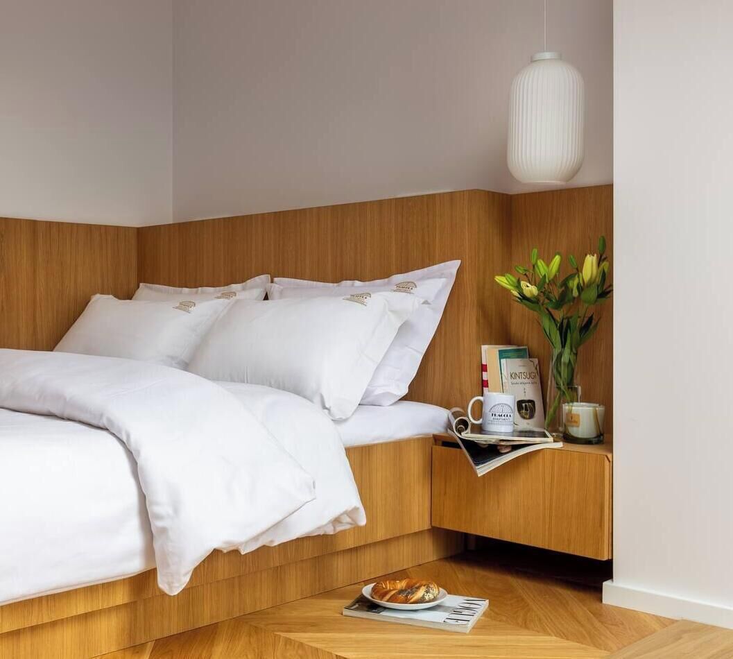 Un lit bien fait, avec du linge de lit blanc, une tête de lit en bois, une table de chevet avec une lampe, un vase rempli de fleurs et un livre.