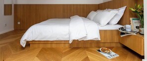 A modern bedroom with a wooden headboard, a neatly made bed, a bedside table with a book and a mug, and a magazine on the floor.