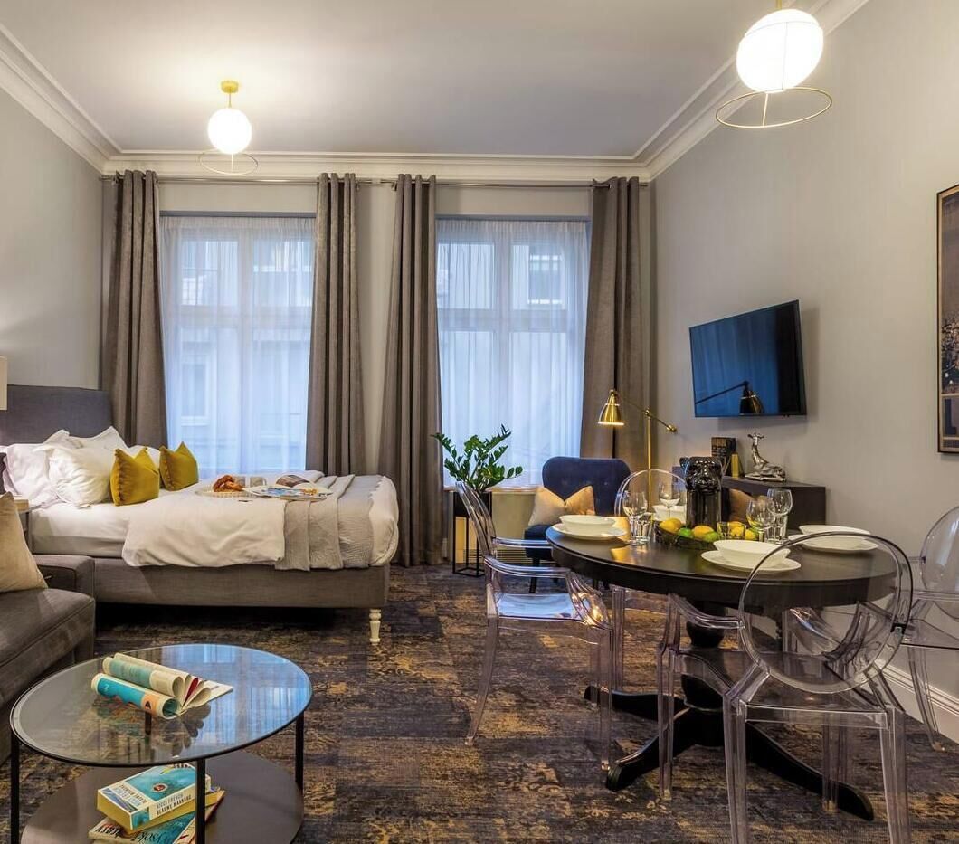 Une chambre d’hôtel comprenant un lit, une table à manger avec des chaises, un téléviseur fixé au mur et une grande fenêtre avec des rideaux.