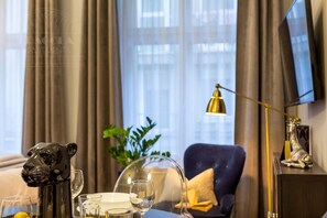 A hotel room with a dining table set with glasses and a decorative item, a blue armchair, a plant, and a TV mounted on the wall.