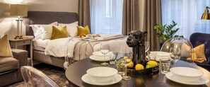 A hotel room with a neatly made bed, a dining table set with plates, glasses, and a fruit bowl.