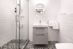 A bathroom with a glass shower enclosure, a white sink with two drawers, a round mirror, and patterned floor tiles.