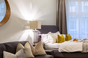 A hotel room with a bed, a bedside table with a lamp, a sofa with cushions, and a window with curtains.