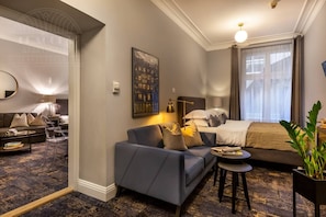 A modern hotel room with a bed, a sofa, a small table, a mirror, and a large window with curtains.