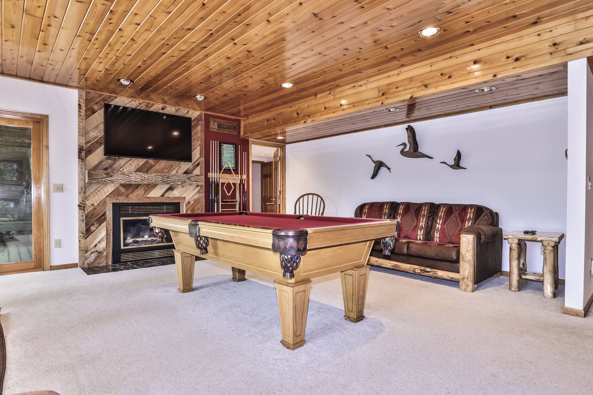 A pool table in a room with wooden ceiling and walls, a fireplace, and a sofa.