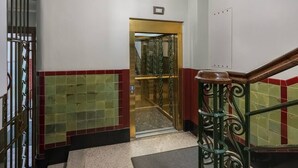 Un couloir avec une grille métallique, un mur recouvert de carreaux rouges et verts, un escalier avec une rampe en bois et un luminaire au plafond.
