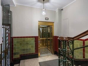 Un pasillo con una reja metálica, un muro de azulejos en rojo y verde, una escalera con barandilla de madera y una lámpara en el techo.