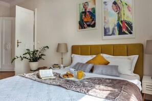 A hotel room with a bed, a bedside table with a lamp, a potted plant, and two paintings on the wall.