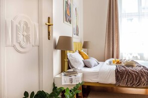 Una habitación de hotel con cama, lámparas de noche, mesita de noche con un libro y una caja, ventana con cortinas y una planta.