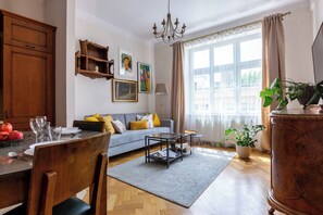 Un salon comprenant un canapé, une table basse, une table à manger avec des chaises, un lustre et une grande fenêtre avec des rideaux.