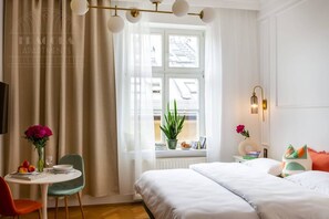 A hotel room with a bed, a table with a chair, a window with curtains, and a plant on the windowsill.