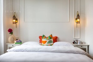 Una cama bien hecha con sábanas blancas, una almohada colorida, un florero con una flor y dos mesitas de noche con lámparas y libros.