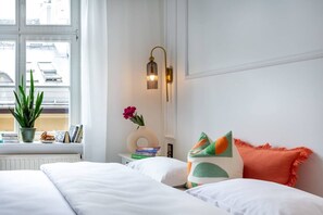 Habitación de hotel con cama, mesita de noche con libros y un florero, ventana con vista a edificios y una lámpara de pared.