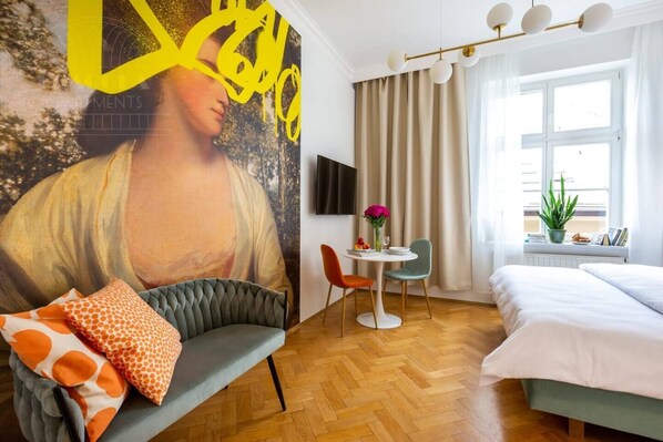 Hotelkamer met een bed, een bank met gestreepte kussens, een eettafel met stoelen, een groot schilderij aan de muur, een televisie en een raam met gordijnen.