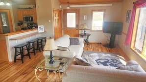 Ein gemütliches Wohnzimmer mit einer Couch, Sesseln, einem Glastisch und einem Fernseher. Es gibt eine Küchenzeile mit Herd, Mikrowelle und Barhockern.
