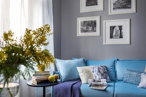 A room with a blue sofa, a small table with books and fruit, and framed pictures on the wall.