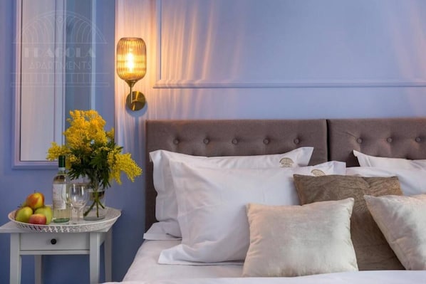 A bedroom with a bed, pillows, a bedside table with fruit and a vase of flowers, and a wall-mounted lamp.