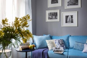A room with a blue sofa, a small table with books and fruit, and framed pictures on the wall.