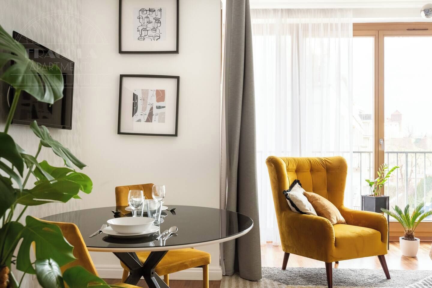 Un coin repas avec une table ronde, des chaises et un fauteuil jaune. Deux œuvres d’art encadrées ornent le mur, ainsi qu’une plante en pot. On peut également apercevoir le paysage à travers une fenêtre.