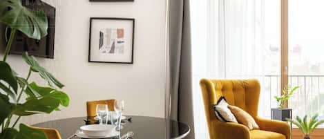 Un coin repas avec une table ronde, des chaises et un fauteuil jaune. Deux œuvres d’art encadrées ornent le mur, ainsi qu’une plante en pot. On peut également apercevoir le paysage à travers une fenêtre.