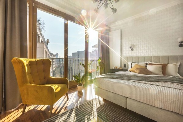 A bedroom with a large bed, a yellow armchair, a potted plant, and a city view through the window. - Sunny studio with balcony (Kraków – Old Town)
