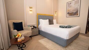 Habitación de hotel con cama, una silla, una mesita con fruta y un cuadro en la pared.