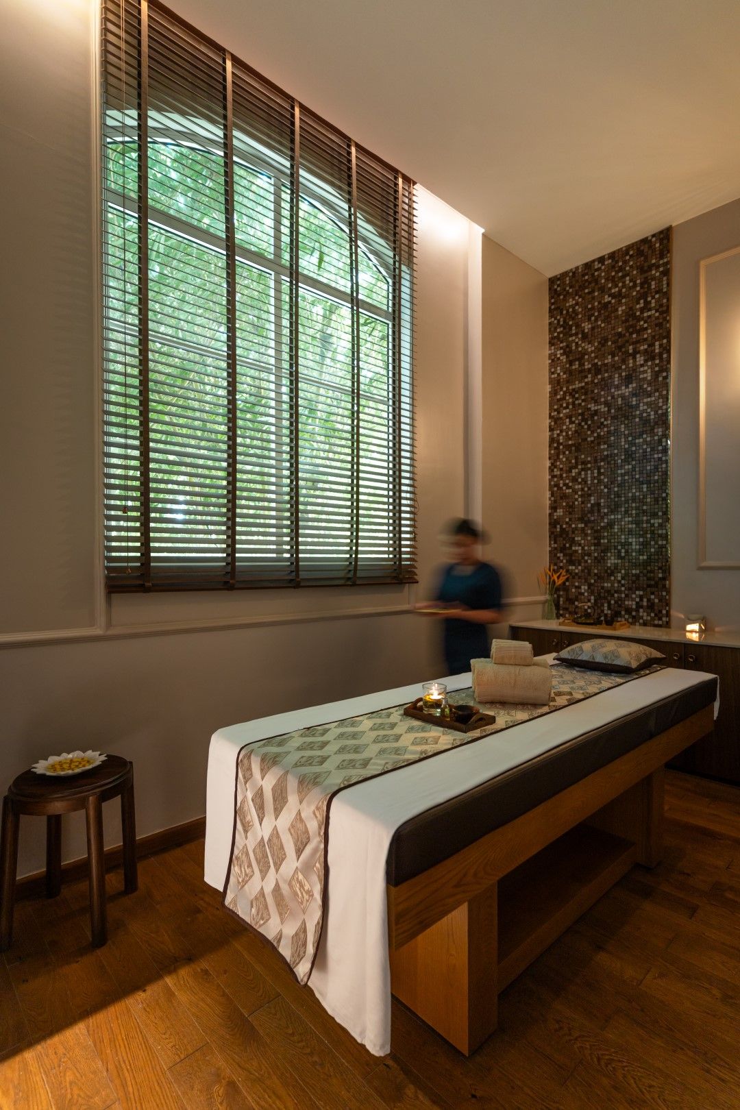 Een kamer met een houten bankje, een tafel met een tafelkleed met patroon, een raam met blinds en een muur met een mozaïekontwerp.