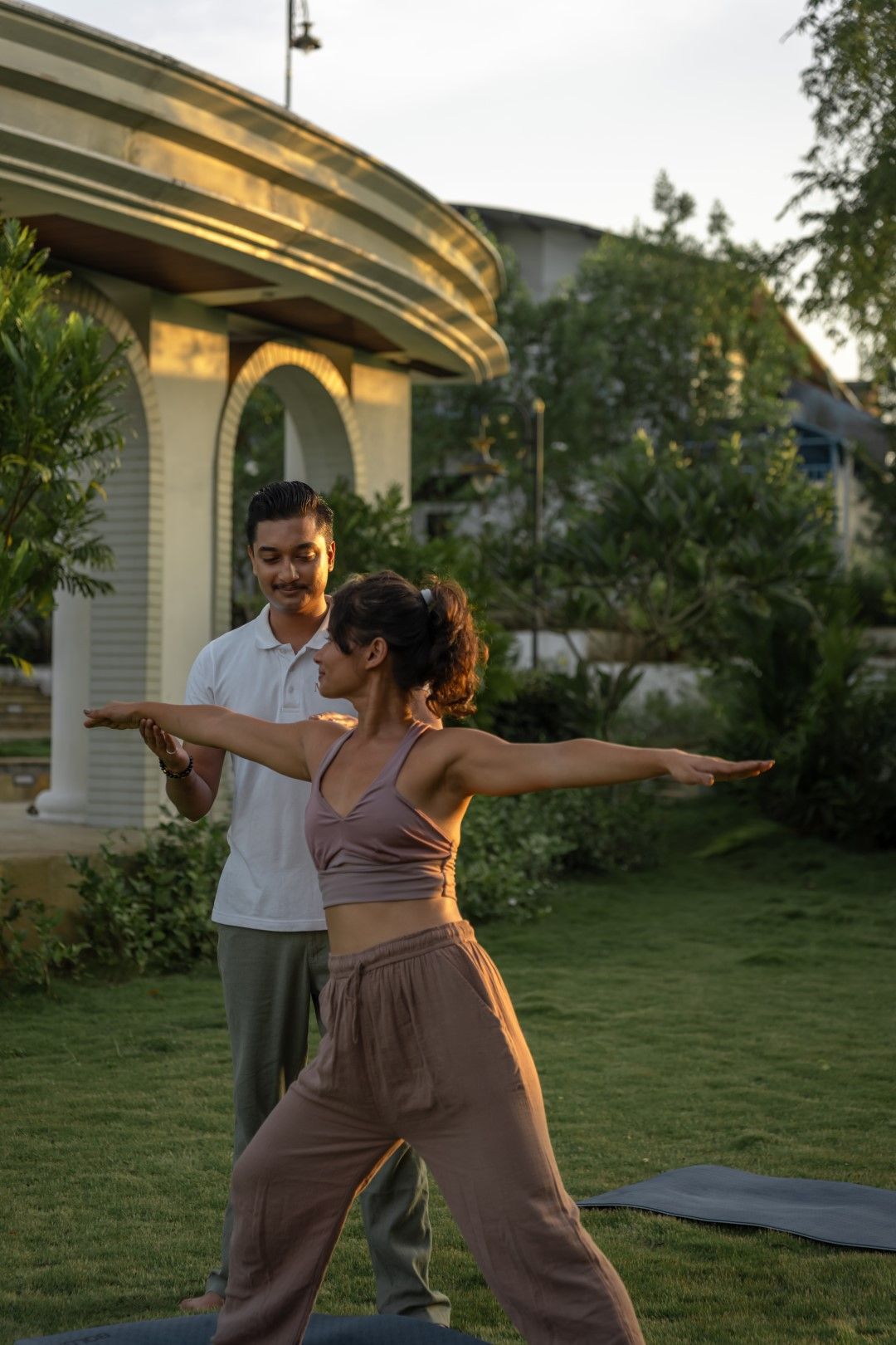 Dos personas practicando yoga al aire libre, con una casa y follaje verde de fondo.