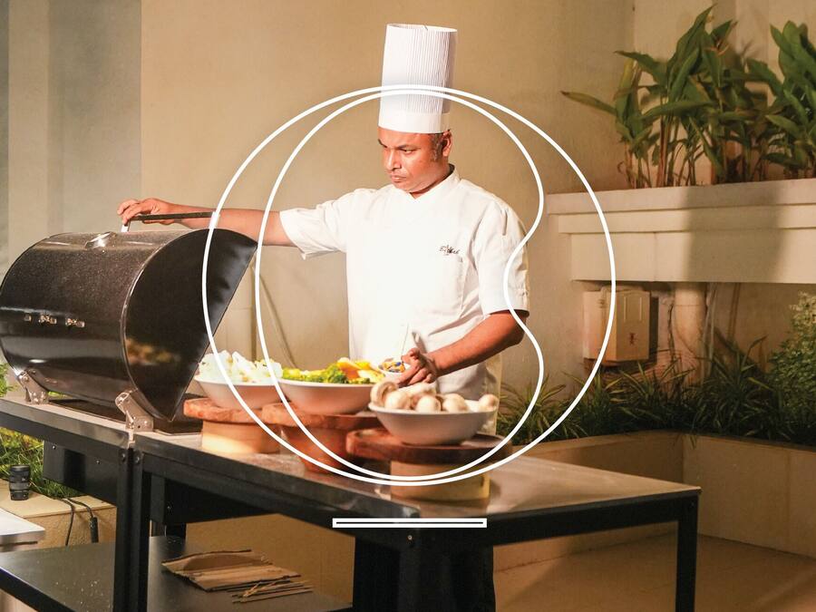A chef in a white uniform preparing food at a table with a grill and bowls of food.