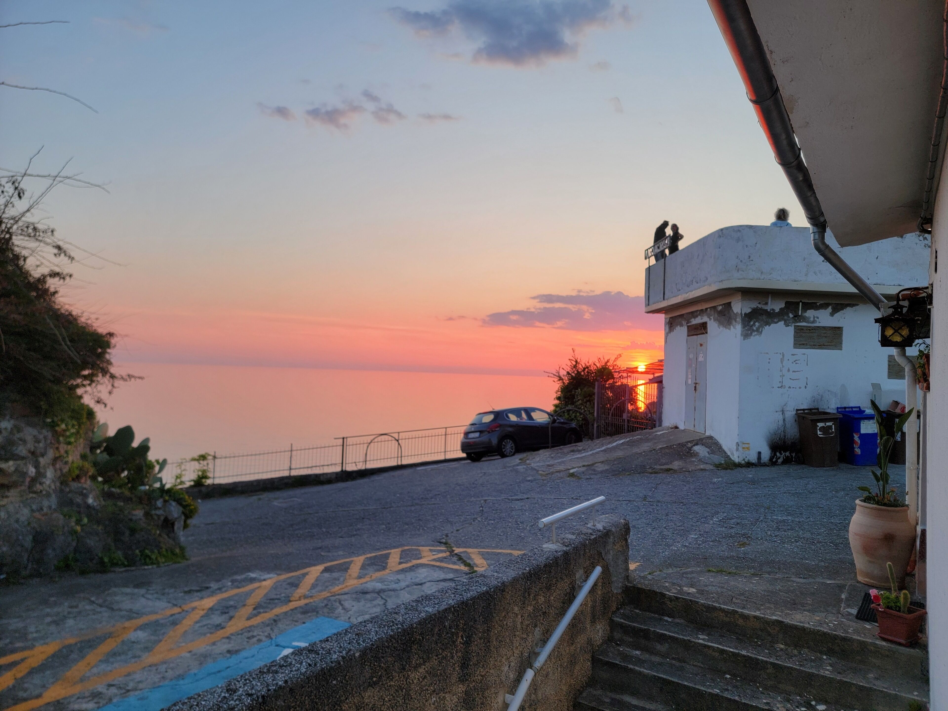 Une route côtière offrant une vue sur le coucher de soleil, une voiture garée et un bâtiment avec des marches menant à l’entrée.