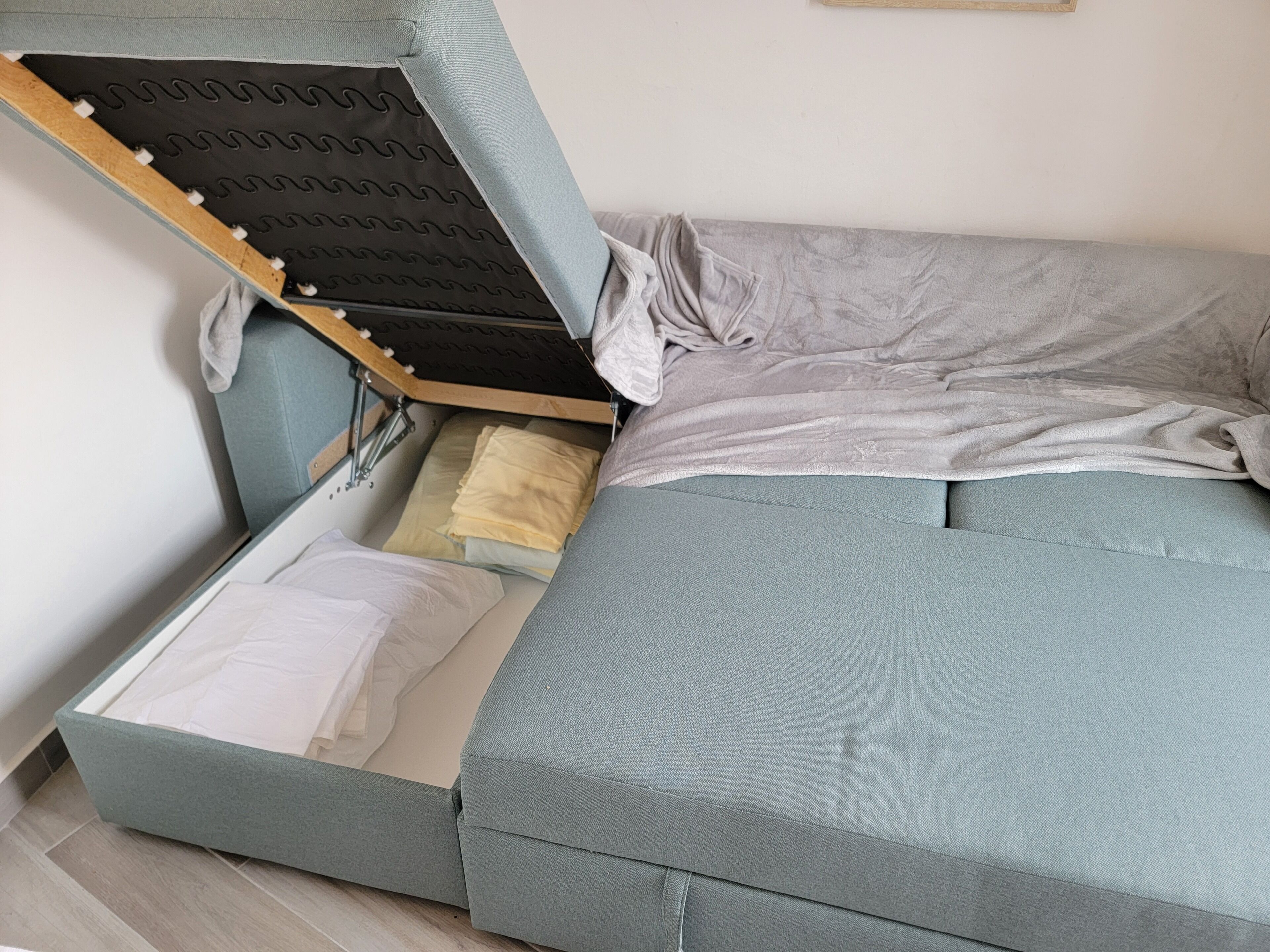 A compact bed with a storage compartment underneath, a grey mattress, and a grey pillow.