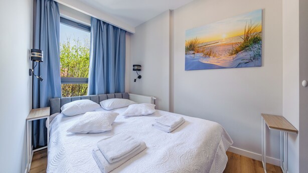 A neatly made bed with white linens and pillows, a painting of a beach scene on the wall, and a window with blue curtains. - Apartament Sun & Snow Kaszubska Bryza ll (Hel)