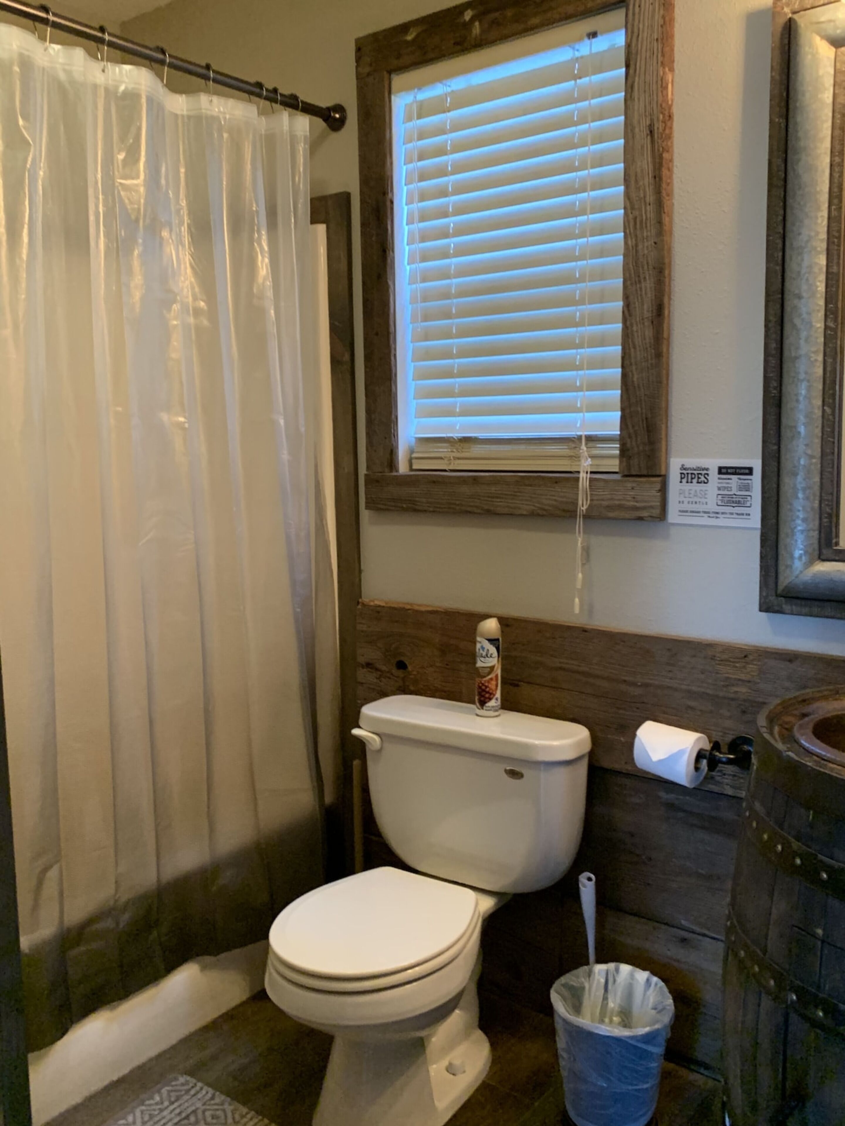 Une salle de bain équipée d’un WC, d’un rideau de douche, d’une fenêtre avec stores, d’un porte-rouleau de papier toilette et d’une poubelle.