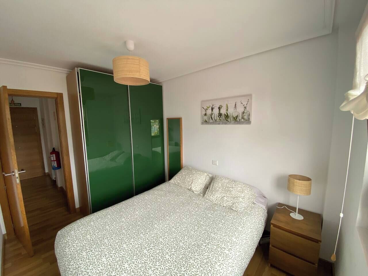 Un dormitorio con una cama, un armario verde, una mesita de noche con lámpara y un cuadro en la pared.