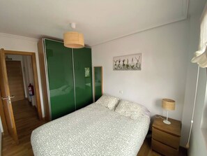 Un dormitorio con una cama, un armario verde, una mesita de noche con lámpara y un cuadro en la pared.