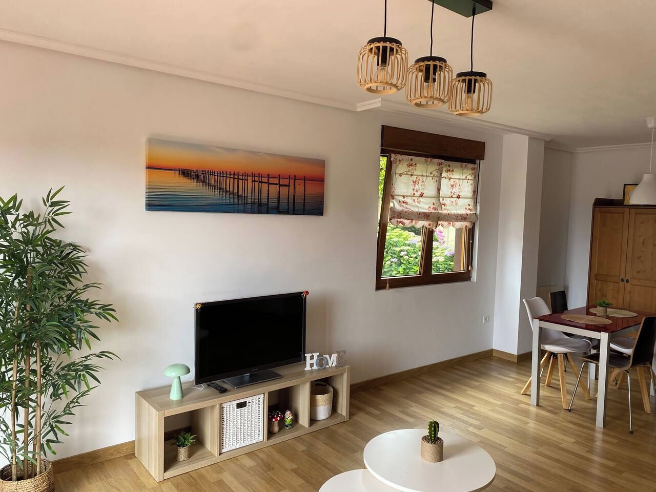 Una sala de estar con televisor de pantalla plana, armarios de madera, una mesa de comedor con sillas, una planta en maceta y un cuadro de una puesta de sol sobre un muelle.
