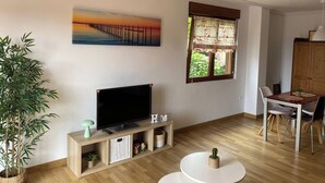 Un salon équipé d’un téléviseur à écran plat, de meubles en bois, d’une table à manger avec des chaises, d’une plante en pot et d’un tableau représentant un coucher de soleil sur une jetée.