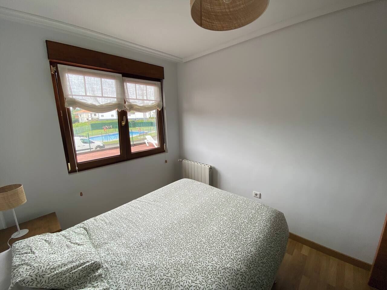 Una habitación con cama, ventana con cortinas, radiador y lámpara sobre una mesita.