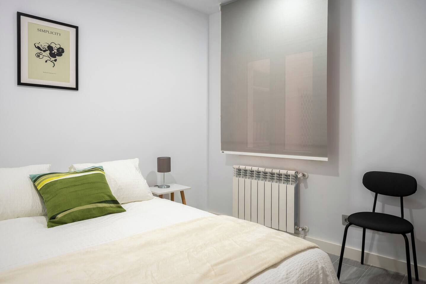 Hotellihuone, jossa on sänky, yöpöytä lampun kanssa, patteri, tuoli ja seinällä oleva kehystetty kuva.