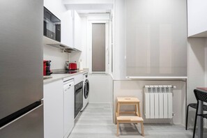 Ett kompakt kök med tvättmaskin, en röd vattenkokare och en liten träpall.