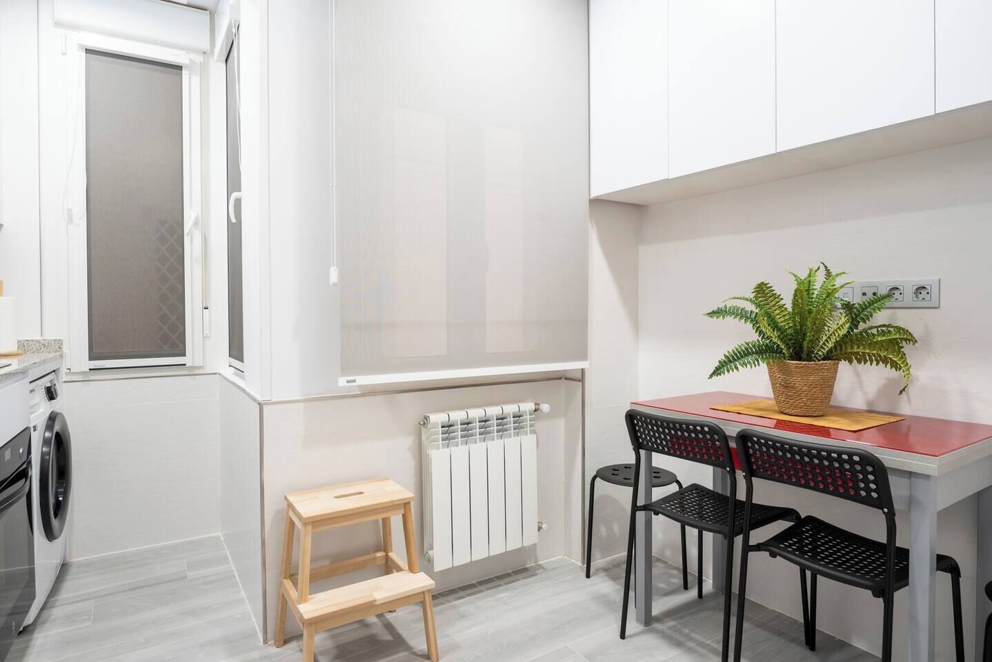 Une petite cuisine équipée d’un lave-linge, d’un radiateur, d’un petit tabouret en bois et d’une table avec deux chaises.
