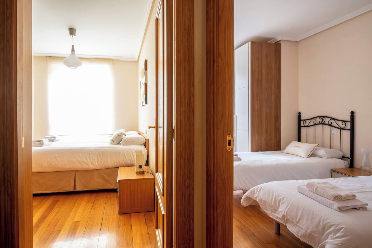 A hotel room with two beds, wooden flooring, a bedside table, and a window with natural light.
