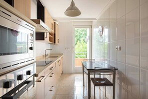 A modern kitchen with a built-in oven, a dining table with chairs, and a view of greenery outside. - Apartamento en Entrambasaguas Centro (Entrambasaguas)